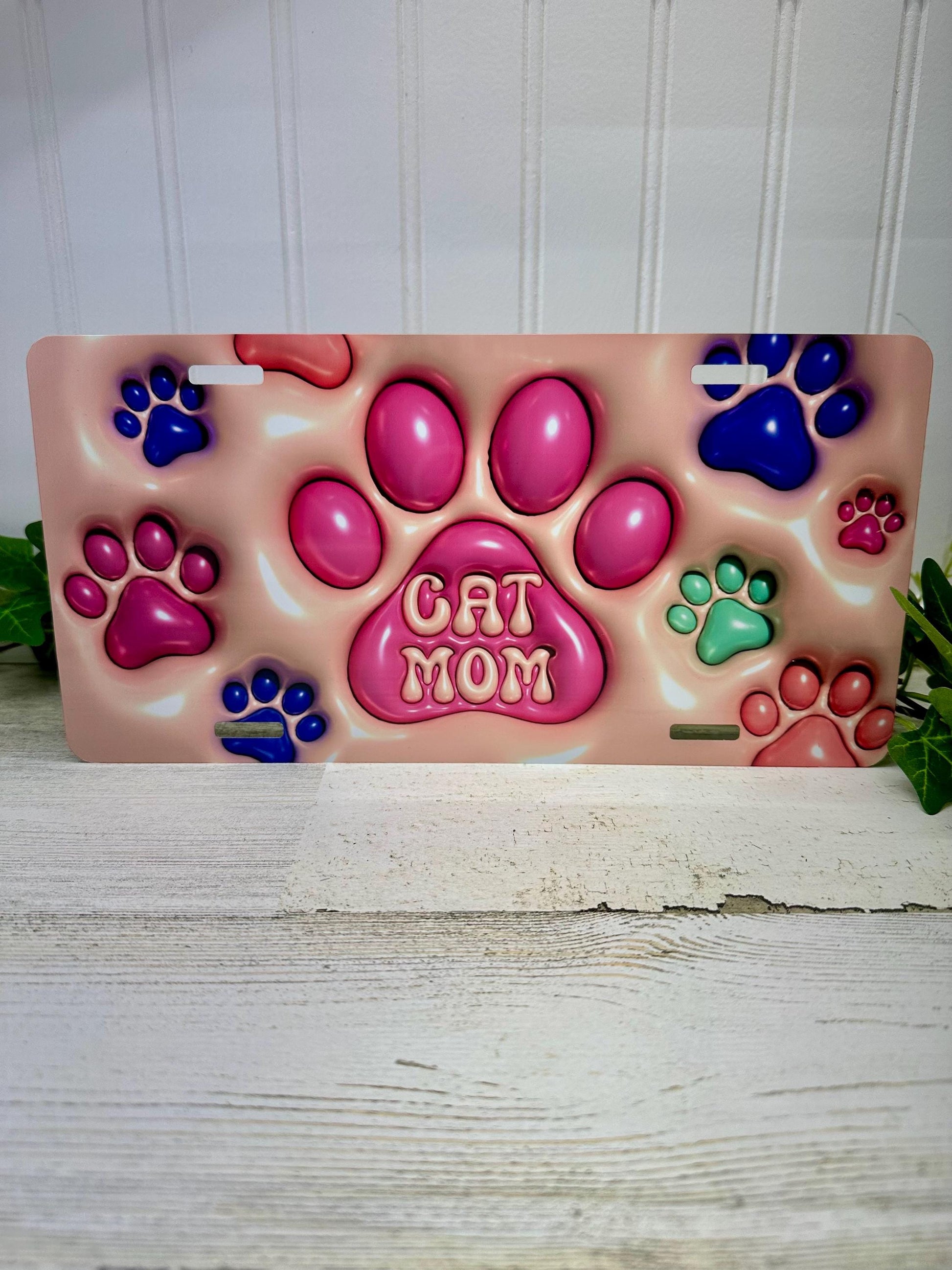 A 3D inflated-style aluminum license plate with a design that includes paw prints and the text 'Cat Mom'.